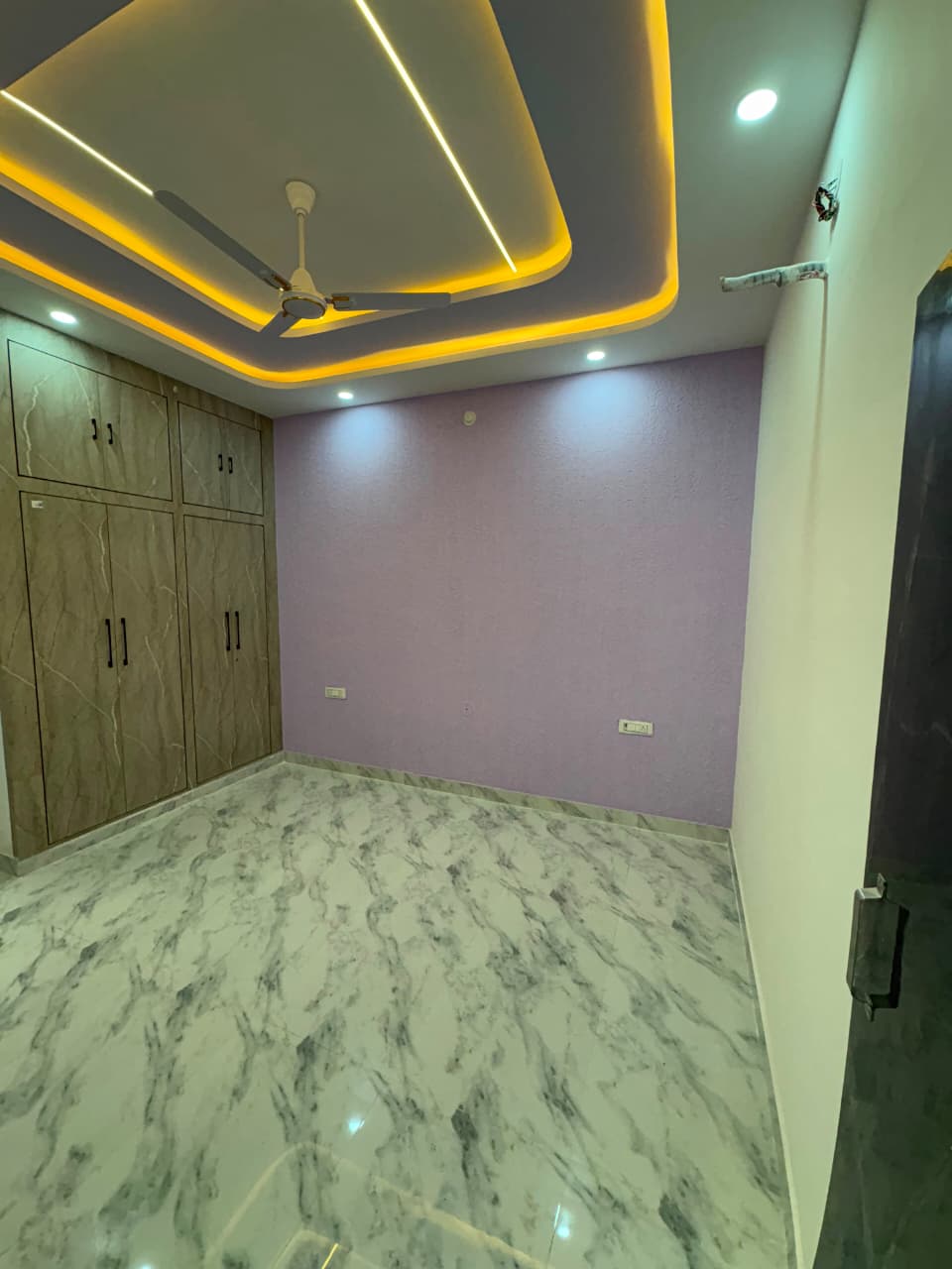 Contemporary bedroom interior with lavender accent wall, warm recessed LED cove ceiling, and built-in laminate wardrobes