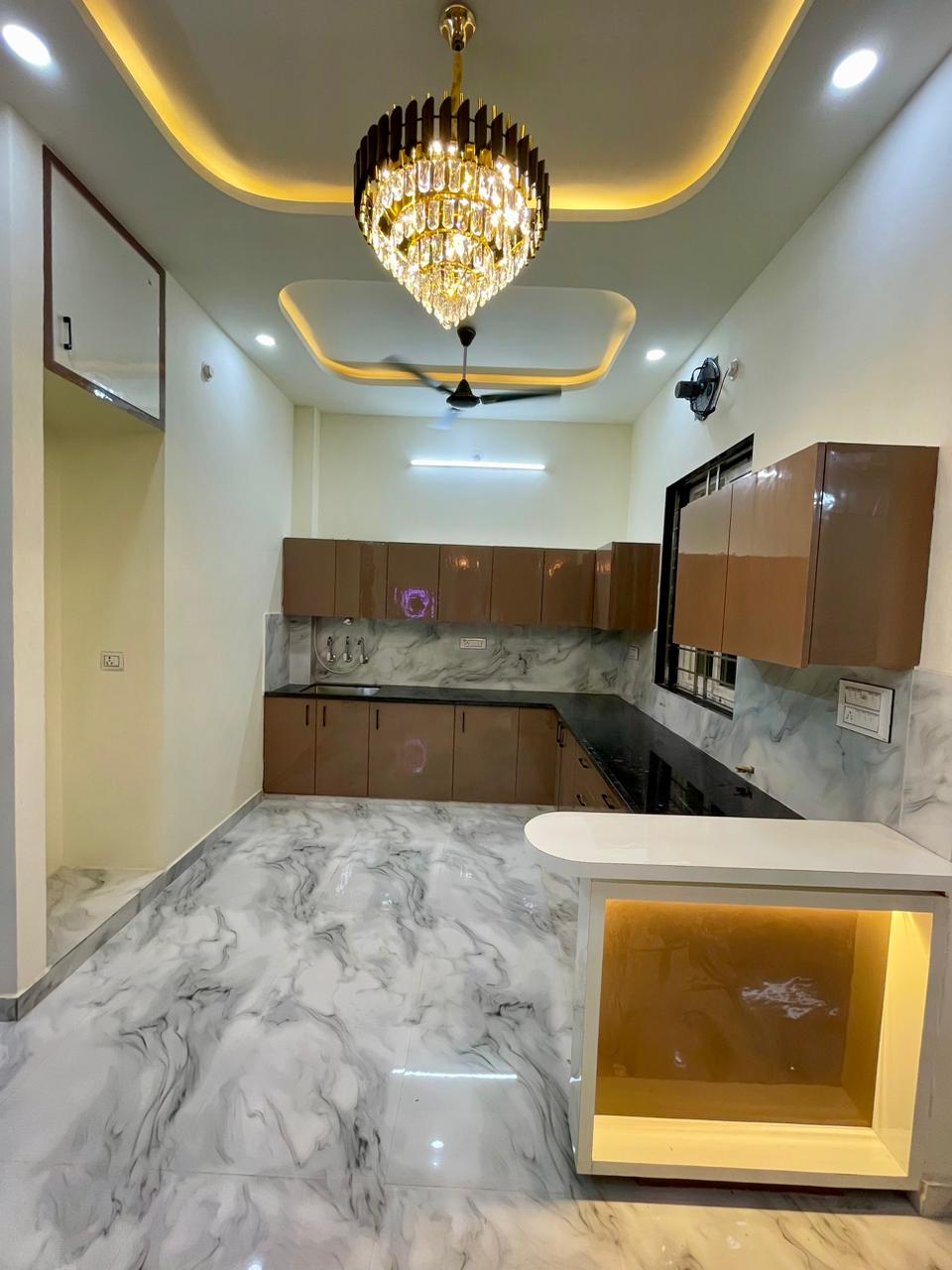 Premium modular kitchen in warm high-gloss finish with illuminated breakfast bar counter, marble backsplash, and crystal ceiling chandelier