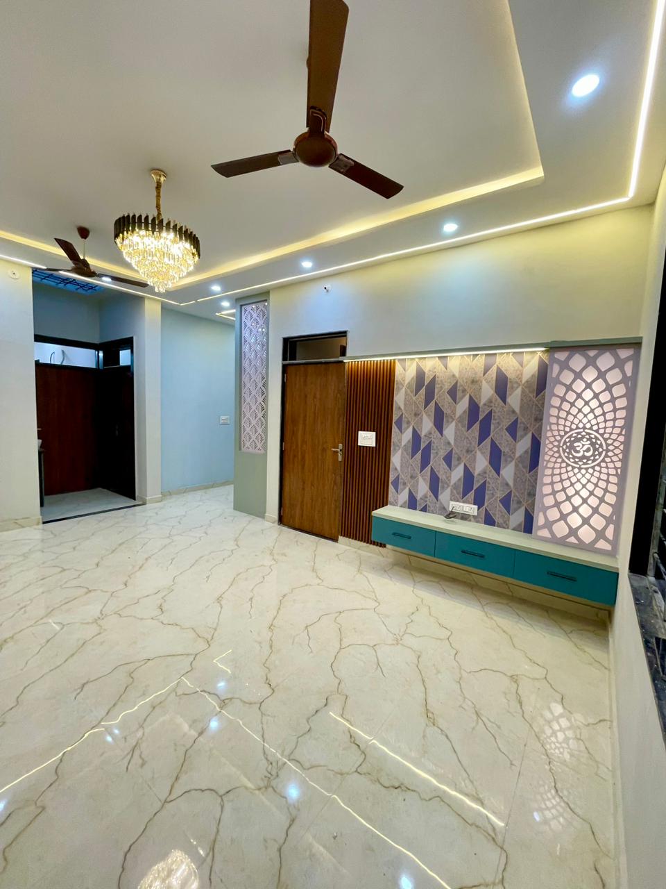Panoramic living hall view showing backlit Om temple jali panel, media console, crystal chandelier, and polished marble flooring